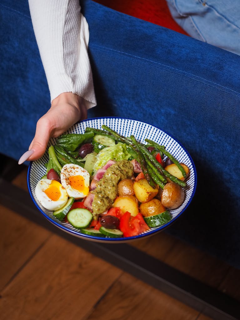 A colorful vegetable salad bowl with eggs and potatoes held by a person in a cozy setting.