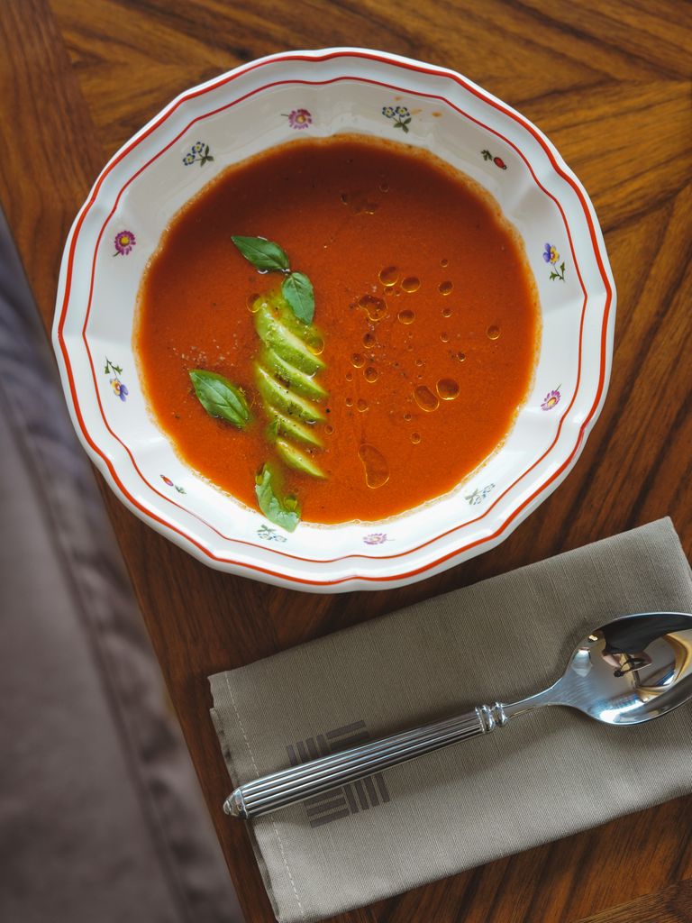 A vibrant bowl of tomato soup topped with basil leaves and sliced avocado on a wooden table.